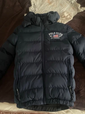 Mörkblå pufferjacka Polo Club - Snygg mörkblå pufferjacka från Polo Club Saint Vincent med huva och dragkedja framtill. Jackan har två sidofickor med dragkedja och tryckt logga på bröstet. Perfekt för kalla vinterdagar och riktigt skön att bära.