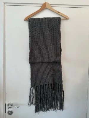 Grå stickad ONLY halsduk med fransar - En lång, stickad halsduk i mörkgrått med bred ribbstickning och långa fransar i ändarna. Perfekt för att hålla värmen under kalla dagar och ger en snygg touch till din outfit.