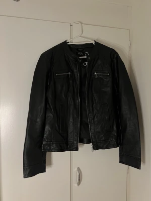 Svart skinnjacka från Only - Snygg svart skinnjacka från Only med två bröstfickor med dragkedja och raka linjer. Jackan har en klassisk bikerstil utan krage och dragkedja framtill. Perfekt för dig som vill ha en stilren och tidlös look.