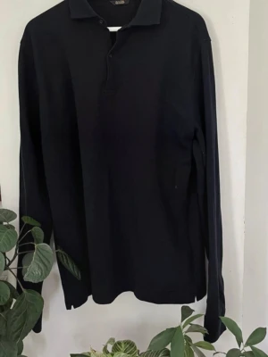 Mörkblå pikétröja i 100% merinoull från Massimo Dutti - Klassisk mörkblå långärmad pikétröja från Massimo Dutti i topp skick. Tröjan har krage, knäppning framtill och är tillverkad i ett mjukt, tunt material som ger en clean och stilren look. Perfekt till jeans eller chinos.