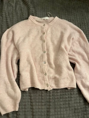 Ljusrosa kofta med knappar - Mysig ljusrosa kofta med rund halsringning och genomgående knäppning framtill. Koftan har långa ärmar och ribbade muddar vid ärmslut och nederkant. Tillverkad i ett mjukt, fluffigt material som känns skönt mot huden.