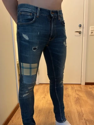 Nudie jeans - Säljer ett par blå skinny jeans med slitningar och en unik rutmönstrad patch på vänster lår. Jeansen har klassisk femficksdesign, dragkedjegylf och kontrastsömmar. Materialet är denim med stretch för skön passform. Perfekta för dig som gillar en trendig och personlig stil.