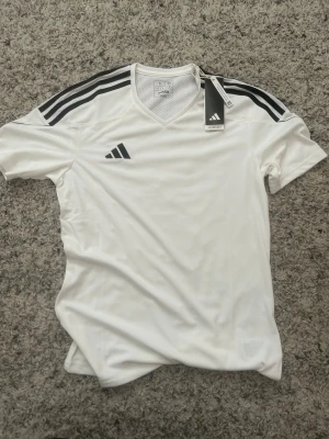 Vit Adidas sporttröja med svarta ränder - Snygg vit sporttröja från Adidas med klassiska svarta ränder på axlarna och Adidas-logga på bröstet. Tröjan har V-ringning och är tillverkad i ett lätt, ventilerande material som passar perfekt till träning eller match.