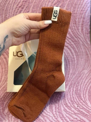 Bruna ribbade strumpor från UGG - Säljer ett par bruna ribbade strumpor från UGG. Strumporna är i en varm brun ton, har en klassisk form och är tillverkade i mjukt material. UGG-logga på kanten ger en snygg detalj. Perfekta för dig som vill ha både stil och komfort.