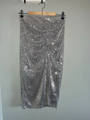 Glittrig silverlångkjol med slits - Säljer en glittrig långkjol i silver med paljetter över hela kjolen. Kjolen har en snygg slits framtill som ger extra edge och är perfekt för dig som vill sticka ut. Rak passform och elastisk midja för skön känsla. Nypris 599kr