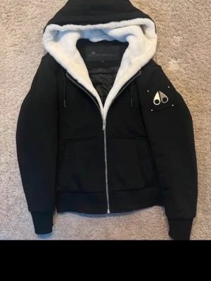 Svart pufferjacka Moose Knuckles - Snygg svart pufferjacka från Moose Knuckles med vit fuskpäls i huvan och quiltat innerfoder. Jackan har dragkedja framtill, två stora fickor och en ikonisk logga på ärmen. Perfekt oversized look och riktigt varm för kalla dagar. Den är ny helt oanvänd 