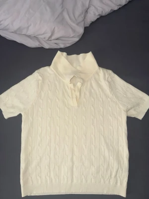 Gul kabelstickad polotopp - Kabelstickad gul polotopp med korta ärmar och bred krage. Toppen har en klassisk kabelstickad struktur och är gjord i ett mjukt material som känns skönt mot huden. Perfekt för en stilren och trendig look.
