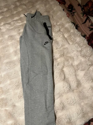 Gråa mjukisbyxor från Nike - Snygga gråa mjukisbyxor från Nike med svart logga på vänster ben och svarta dragsnören i midjan. Byxorna har en dragkedjeförsedd ficka på sidan och är tillverkade i mjukt sweatshirtmaterial. Perfekta för chill eller träning.