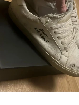 Saint Laurent vita låga sneakers - Vita sneakers från Saint Laurent med handskriven logga på sidan. Skorna har slitna detaljer, rund tå och klassisk snörning. Tillverkade i canvas med råa kanter för en edgy look. Perfekta för dig som gillar streetstyle och designer vibes.