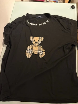 Svart Burberry t-shirt med nalle - Svart t-shirt från Burberry med klassiskt rutigt nallemotiv på bröstet och Burberry-tryck runt halsringningen. T-shirten har korta ärmar och är gjord i mjuk bomull. Perfekt för dig som vill ha en ikonisk och lekfull stil.