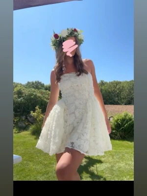Vit spetsklänning med volang - Supersöt vit kort klänning med blommig spets och volangkant. Klänningen är axelbandslös och har en utsvängd kjol. Perfekt till sommaren