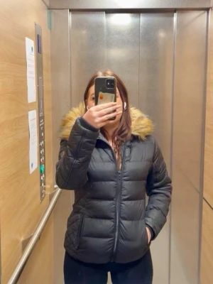 Svart pufferjacka med fuskpäls - Snygg svart pufferjacka med dragkedja och huva med beige fuskpäls. Jackan har en klassisk quiltad design och är perfekt för kalla dagar. Fodrad och med fickor på sidorna. Passar dig som vill ha en varm och trendig vinterjacka.