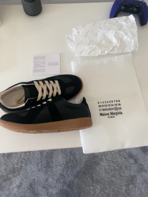 Maison Margiela svarta sneakers - Maison Margiela sneakers i svart med beige snörning och klassisk gummisula. Skorna har en stilren och tidlös design med materialmix av mocka och läder. Perfekta för dig som gillar minimalistisk och exklusiv stil.