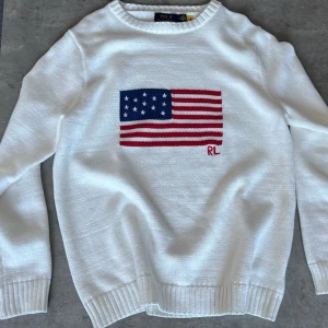 Vit stickad tröja Polo Ralph Lauren - Vit stickad tröja från Polo Ralph Lauren med amerikansk flagga och RL-broderi på bröstet. Tröjan har rund halsringning, ribbade muddar och nederkant. Perfekt för dig som gillar klassisk och stilren design.