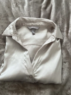 Beige sidenliknande skjorta H&M - Stilren beige skjorta från H&M i ett glansigt, sidenliknande material. Klassisk krage, knäppning framtill och långa ärmar. Perfekt för dig som gillar en enkel men elegant look. Har använt den flitigt men tagit hand om hyfsat.