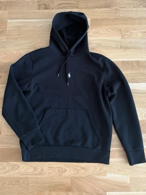 Svart hoodie från Ralph Lauren - Snygg svart hoodie från Ralph Lauren med klassisk huva och dragsnören. På bröstet sitter den ikoniska vita Polo-loggan. Hoodien har en stor magficka och ribbade muddar vid ärmar och nederkant. Perfekt för en avslappnad och stilren look.