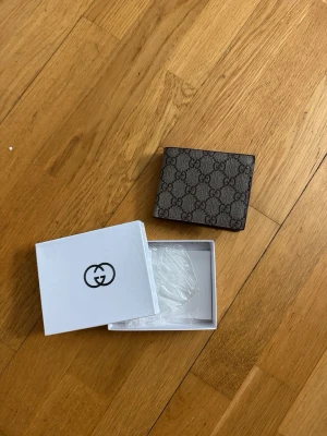  G plånbok - Säljer en brun plånbok från Gucci i klassiskt GG Supreme-mönster. Plånboken är i canvas med bruna läderdetaljer och har flera fack för kort och sedlar. Kommer med original ask. Perfekt för dig som gillar lyxiga accessoarer.