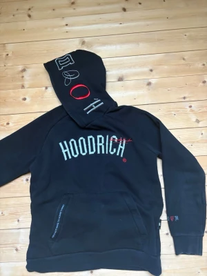 Svart HOODRICH hoodie med brodyr - Svart hoodie från HOODRICH med stor broderad logga på bröstet och detaljer i vitt och rött på huvan och ärmen. Stora fickor framtill och tryck på ryggen. Perfekt för dig som gillar streetwear och vill sticka ut.