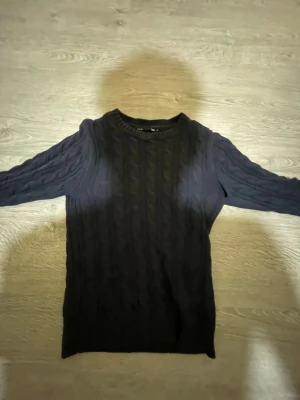 Svart kabelstickad tröja 157 - Kabelstickad svart tröja från 157 med rund halsringning och långa ärmar. Tröjan har ett klassiskt flätmönster och ribbade muddar vid hals, ärmslut och nederkant. Perfekt för lager på lager under kyliga dagar.