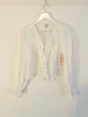 Nelly Stunning Flounce Blouse - Söt vit blus från Nelly med volangdetaljer framtill och knytband vid ringningen. Blusen har långa ärmar med knyt vid ärmsluten och fina veckade detaljer upptill. Tillverkad i ett tunt, lätt tyg som jag kan tänka mig är så skönt på sommaren. Toppen har alla taggar kvar, ordinarie pris 399.