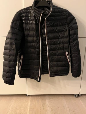 Svart pufferjacka med randig dragkedja - Säljer en svart pufferjacka med quiltad design och hög krage. Jackan har två fickor med dragkedja som har snygga ränder i vitt, rött och blått. Materialet är glansigt och lätt, perfekt för lite kallare vårdagar. Passar dig som vill ha en stilren och sportig look. Jackan är i mycket bra skick förutom en litet hål (bild 3) som man bara ser från extremt nära håll därav det låga priset. Självklart så är jackan äkta. Storlek S