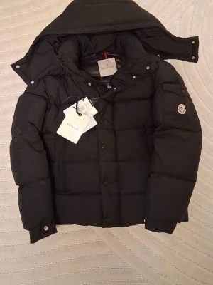 Svart dunjacka från Moncler - Snygg svart dunjacka från Moncler med avtagbar huva och klassisk logotyp på ärmen. Jackan har dragkedja och knappar framtill, samt praktiska fickor och en innerficka med serietidningsmönster. Perfekt för kalla vinterdagar och riktigt varm.