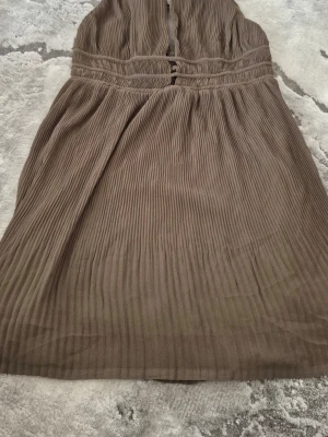 Brun plisserad klänning - Snygg brun klänning med plisserad design och markerad midja. Klänningen har en mjuk och luftig känsla, perfekt för dig som gillar en avslappnad men ändå stilren look. Axelbandslös modell som ger en modern vibe.