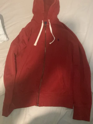 Röd hoodie med dragkedja och fickor - Röd hoodie med hel dragkedja framtill, vita snören i huvan och två stora fickor. Klassisk och enkel design, perfekt för en avslappnad stil. Liten broderad logga på bröstet.