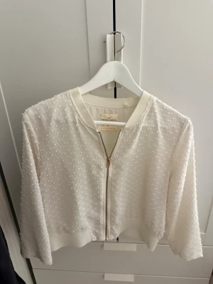 Vit prickig zip-kofta - Söt vit kofta med dragkedja från Holly & Whyte. Koftan har ett diskret prickigt strukturmönster, ribbade muddar och är tillverkad i ett tunt, lätt material. Perfekt att slänga över en topp för en fräsch look.