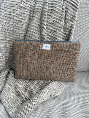 Beige clutch från Emelo i ullmix - Stilren beige clutch från Emelo i grovstickad ullmix. Väskan har en enkel rektangulär form och stängs med dragkedja upptill. Perfekt för att bära småsaker eller använda som necessär. Mått ca 29x19cm
