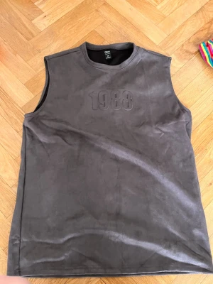 Mörkgrå ärmlös topp med ”1988”  - Cool mörkgrå ärmlös topp med präglad text '1988' på bröstet. Materialet har en lätt mockakänsla och toppen har en clean, enkel design som passar perfekt till jeans eller shorts. Skriv gärna för mått! 💕