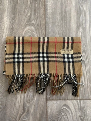 Beige rutig halsduk från Burberry - Klassisk beige halsduk från Burberry i 100% lammull med det ikoniska rutmönstret i svart, vitt och rött. Halsduken har fransar i ändarna och en snygg Burberry-label. Perfekt accessoar för att lyfta din outfit.