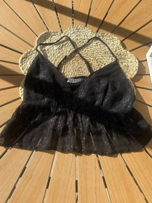 Glittrig svart festopp från NA-KD - Säljer en glittrig svart festtopp från NA-KD med tunna axelband och v-ringad hals. Toppen har en lätt volang nedtill och är croppad i modellen. Perfekt för dig som vill ha något extra till outfiten!🖤