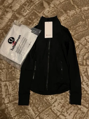 Svart jacka från Lululemon - Snygg svart jacka från Lululemon med hög krage och hel dragkedja framtill. Jackan har två fickor med dragkedja och långa ärmar med tumhål. Perfekt för en sportig och stilren look.