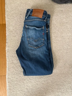 Blå jeans från Replay - Säljer ett par klassiska blå jeans från Replay med snygg tvätt och raka ben. Jeansen har fem fickor, läderpatch med logga bak och coola slitningar på framsidan. Perfekta för en avslappnad och trendig look.