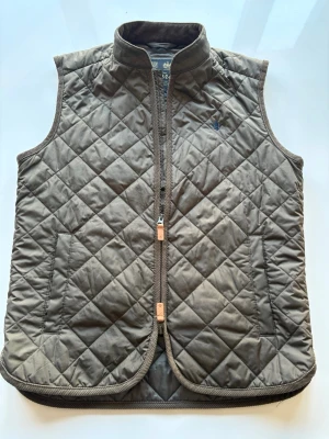 Brun quiltad dunväst från Morris längs ungefär från 165-175 - Snygg brun dunväst från Morris med quiltat mönster och ståkrage. Västen har två fickor framtill och dragkedja med läderdetaljer. Perfekt att slänga över en hoodie eller tröja för extra värme. Diskret morris logga broderad på bröstet. Org pris 1999 