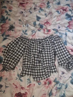 Svartvit rutig offshoulder blus - Trendig svartvit rutig blus med offshoulder och puffiga trekvartsärmar. Blusen har volangdetaljer vid ärmslut och nederkant samt en smockad resår upptill för en snygg passform. Perfekt för dig som vill ha en söt och stilren topp med lite extra attityd. (Säljer pga att det inte är mun storlek)