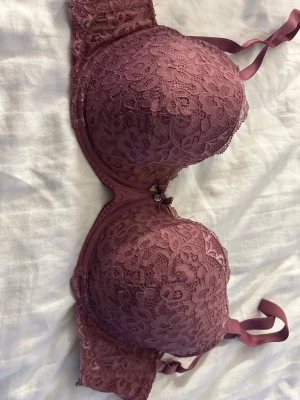 Rosa push-up bh med spetsdetaljer - Säljer en rosa push-up bh från Lindex med vacker blommig spets över hela kuporna och sidorna. Bh:n har en liten rosett och en silverfärgad detalj framtill mellan kuporna. Axelbanden är justerbara och materialet känns mjukt och följsamt.