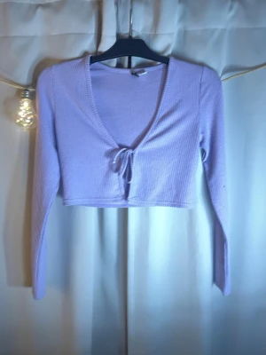 Ljuslila croppad ribbad topp - Trendig ljuslila croppad topp med långa ärmar och v-ringning. Toppen har en söt knytning framtill och är gjord i ribbad bomullsmix som ger en skön och stretchig passform. Perfekt att styla med höga jeans eller kjol.