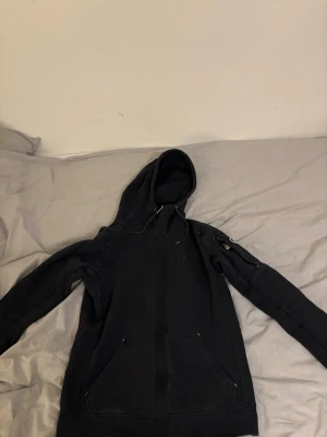 Svart hoodie från Pelle P med patch - Snygg svart hoodie från Pelle P med dragkedja, stor huva och fickor framtill. På ärmen sitter en rund patch med Pelle P-loggan och svensk flagga. Hoodien har ribbade muddar och detaljerade sömmar för en sportig look.