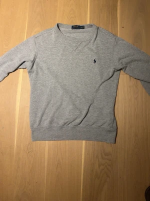 Grå sweatshirt från Polo Ralph Lauren - Klassisk grå sweatshirt från Polo Ralph Lauren med rund halsringning och den ikoniska mörkblå logotypen broderad på bröstet. Tröjan har ribbade muddar vid ärmslut och nederkant. Tillverkad i mjuk bomullsblandning för en skön känsla.