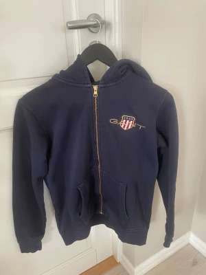 Mörkblå hoodie från Gant - Mörkblå hoodie från Gant med dragkedja framtill och fickor på magen. Klassisk logga och broderad sköld på bröstet i rött, vitt och blått. Tillverkad i mjukt bomullsmaterial och har en skön huva. Perfekt för en avslappnad och sportig stil.