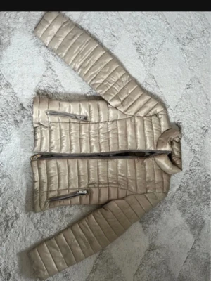 Guld lätt dunjacka med dragkedjor - Snygg guld/beige dunjacka med quiltad design och två dragkedjeförsedda fickor framtill. Jackan har hög krage och smal passform som ger en stilren look. Perfekt för kalla vinterdagar och enkel att matcha med olika outfits.