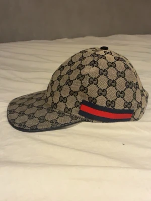 Gucci beige keps med GG-mönster - Säljer en beige keps från Gucci med det klassiska GG-monogrammet över hela kepsen. På sidan finns en ikonisk röd och marinblå rand. Kepsen har böjd skärm och justerbar rem bak. Materialet är canvas med detaljer i tyg. Perfekt för dig som vill ha en lyxig och trendig accessoar.