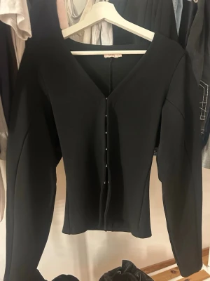 Svart figurnära kofta med knappar - Stilren svart kofta med figurnära passform och djup V-ringning. Koftan har långa ärmar och knäpps framtill. Perfekt att matcha med både jeans och kjol för en trendig look. Använd 1 gång!!