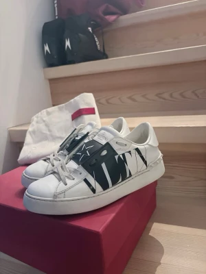 Valentino vita sneakers med svart detalj - Snygga vita sneakers från Valentino Garavani med svart band och stor VLTN-logga på sidan. Skorna har vita snören, rund tå och är tillverkade i läder. Kommer med original dustbag och röd låda. Perfekta för dig som gillar exklusiv streetstyle.        Storlek: 36 2/3 Originalkartong och dustbag ingår.
