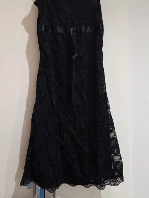 Svart spetsklänning med satinband - Elegant svart långklänning i spets med blommigt mönster och ett dekorativt satinband i midjan. Klänningen har en klassisk siluett och fina spetsdetaljer som ger en lyxig känsla. Perfekt för dig som vill ha en stilren och tidlös look.