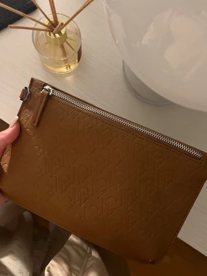 Brun kuvertväska  - Snygg brun kuvertväska från Carin Wester i präglat konstläder med logotypmönster över hela väskan. Väskan har silverfärgad dragkedja och minimalistisk design som gör den enkel att matcha till olika outfits.