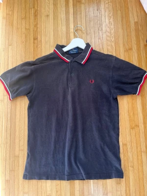 Svart Fred Perry piké med röd/vit kant - Klassisk svart pikétröja från Fred Perry med röd och vit rand på krage och ärmslut.  Tillverkad i bomull. Skick enligt bild