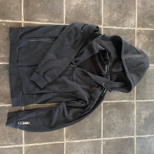 Svart hoodie från Sail Racing - Svart hoodie från Sail Racing med dragkedja framtill och meshfoder i huvan. Snygg logga på ärmen och diskreta detaljer längs dragkedjan. Tillverkad i mjukt material som känns bekvämt och sportigt. Perfekt för dig som gillar en clean och stilren look. Pris ej hugget i sten, postar inon 1-2 dagar.